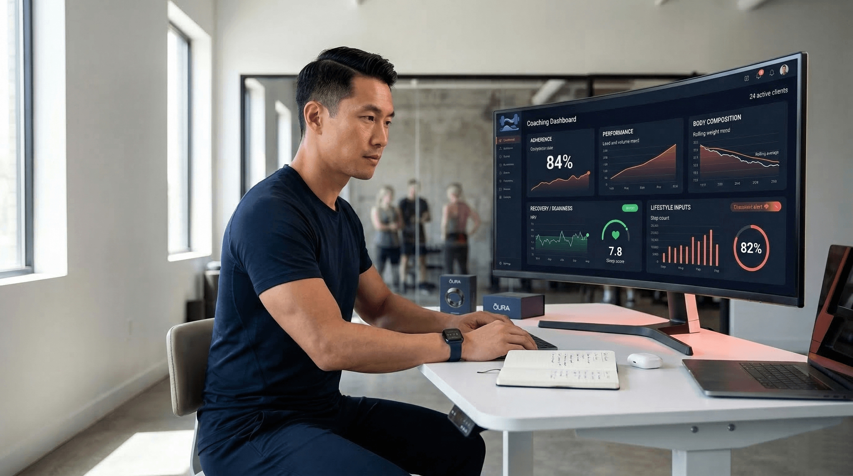 East Asian male personal trainer reviewing a five-metric client dashboard on a large dark-mode monitor showing adherence rates, performance trends, and HRV data for 24 active clients